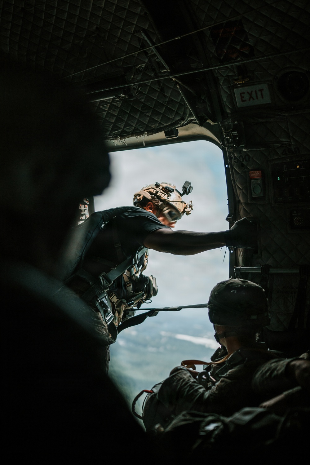 The image shows military personnel inside what appears to be a military helicopter or transport aircraft, with an open side door revealing sky and terrain below. At least two individuals in tactical gear and helmets equipped with night vision mounts are visible, with one leaning toward the open door. The scene suggests an airborne operation, reconnaissance, or insertion/extraction mission in progress.