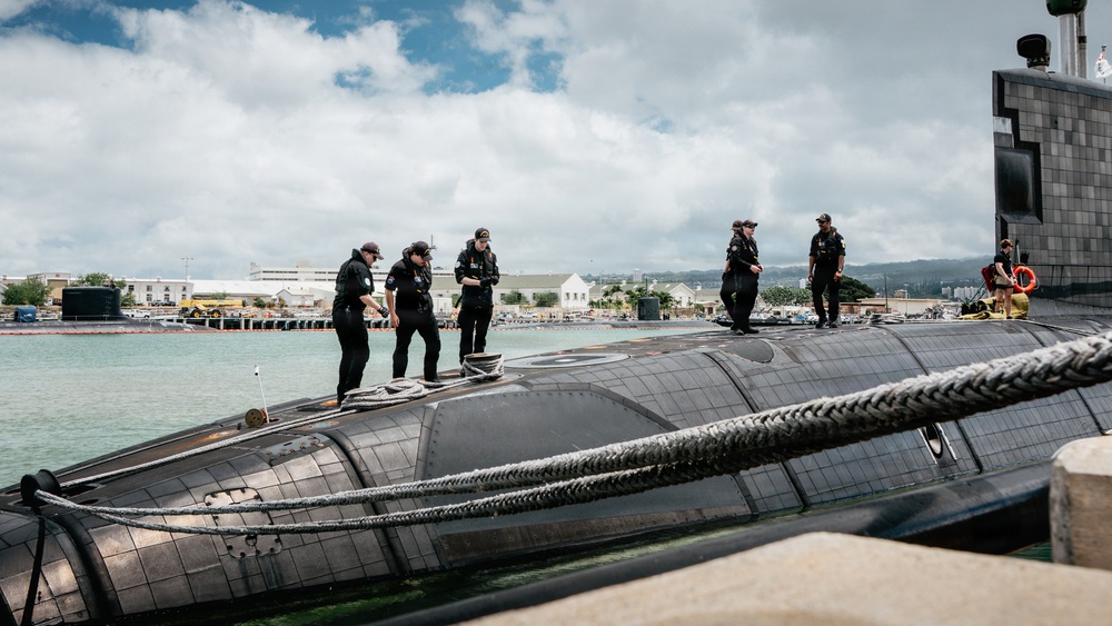A group of naval personnel dressed in black uniforms are standing on the hull of a submarine moored at a naval base. The submarine appears to be docked with mooring lines visible, and a naval facility with buildings and other vessels can be seen in the background. The setting appears to be a tropical or subtropical port environment based on the vegetation and architecture visible.