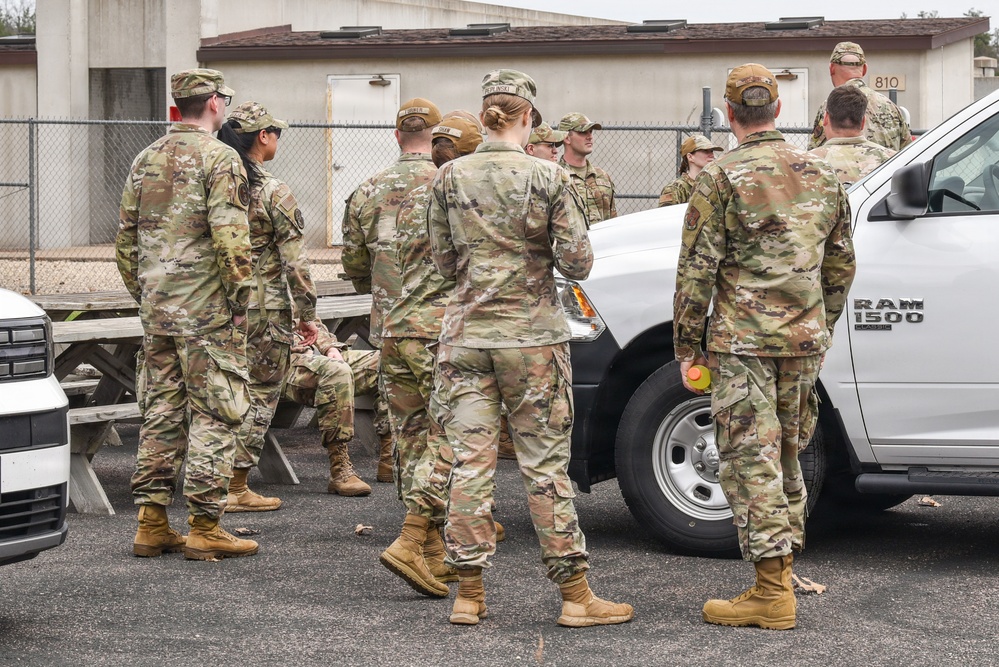 A group of military personnel in OCP (Operational Camouflage Pattern) uniforms are gathered in a parking area near a white Ram 1500 pickup truck. The setting appears to be a military installation, with a numbered building (810) and chain-link fencing visible in the background. The group appears to be receiving a briefing or conducting a formation.