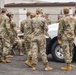 A group of military personnel in OCP (Operational Camouflage Pattern) uniforms are gathered in a parking area near a white Ram 1500 pickup truck. The setting appears to be a military installation, with a numbered building (810) and chain-link fencing visible in the background. The group appears to be receiving a briefing or conducting a formation.