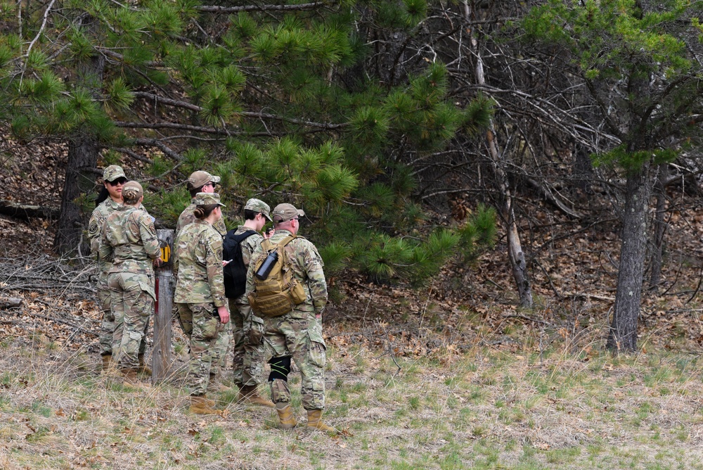 A group of approximately five to six military personnel in OCP (Operational Camouflage Pattern) uniforms are gathered near a wooden post at the edge of a forested area. They appear to be reviewing materials or receiving a briefing in an outdoor training environment. The setting suggests a field exercise or land navigation activity.