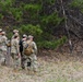 A group of approximately five to six military personnel in OCP (Operational Camouflage Pattern) uniforms are gathered near a wooden post at the edge of a forested area. They appear to be reviewing materials or receiving a briefing in an outdoor training environment. The setting suggests a field exercise or land navigation activity.