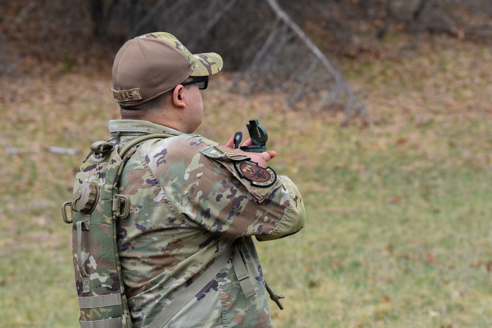 A military service member in MultiCam/OCP camouflage uniform is photographed from behind while using a lensatic compass, likely conducting land navigation training. The individual is wearing a tactical backpack, protective eyewear, and a camouflage cap with a visible name tape reading 'ARINAS'. A unit patch is visible on the left shoulder of the uniform.