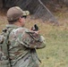 A military service member in MultiCam/OCP camouflage uniform is photographed from behind while using a lensatic compass, likely conducting land navigation training. The individual is wearing a tactical backpack, protective eyewear, and a camouflage cap with a visible name tape reading 'ARINAS'. A unit patch is visible on the left shoulder of the uniform.