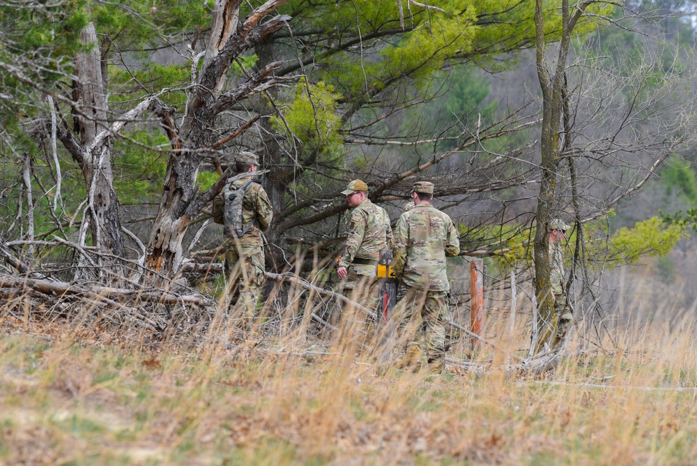 Four military personnel in camouflage uniforms are moving through a wooded, open field environment. They appear to be conducting a field training exercise or patrol, navigating through sparse trees and dry grassland. One individual is carrying a yellow-marked item, possibly training equipment or a marker.