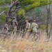 Four military personnel in camouflage uniforms are moving through a wooded, open field environment. They appear to be conducting a field training exercise or patrol, navigating through sparse trees and dry grassland. One individual is carrying a yellow-marked item, possibly training equipment or a marker.