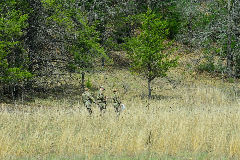 Three individuals in military camouflage uniforms are standing in an open grassy field, appearing to consult a document or map. They are positioned in the middle distance against a backdrop of mixed deciduous and coniferous trees on a hillside. The scene suggests a field training exercise or land navigation activity.