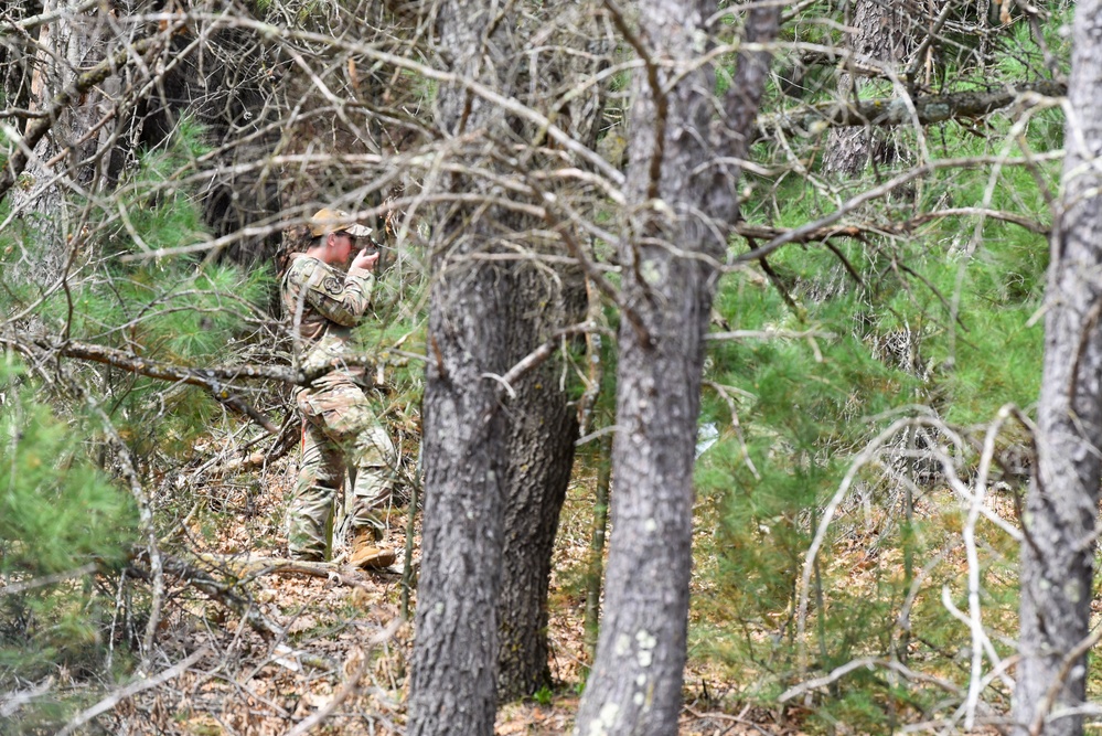A person in military camouflage uniform and cap is positioned among trees in a wooded area, using binoculars or a similar optical device to observe something in the distance. The individual appears to be conducting reconnaissance or surveillance from a concealed position within the tree line. The forest setting provides natural cover with pine and bare-branched trees surrounding the subject.