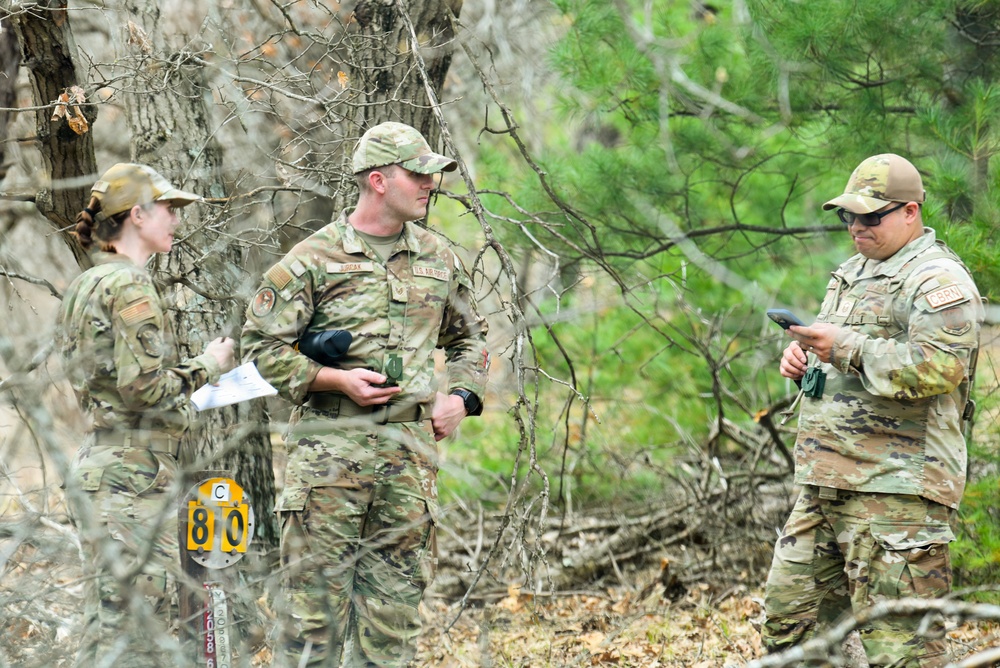 Three U.S. Air Force personnel in OCP (Operational Camouflage Pattern) uniforms are gathered in a wooded area, appearing to conduct a land navigation or field training exercise. One individual is holding papers/documents, the center figure holds binoculars, and the right figure is examining a handheld device. A yellow marker sign with the letter 'C' and number '80' is visible in the foreground.