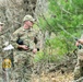 Three U.S. Air Force personnel in OCP (Operational Camouflage Pattern) uniforms are gathered in a wooded area, appearing to conduct a land navigation or field training exercise. One individual is holding papers/documents, the center figure holds binoculars, and the right figure is examining a handheld device. A yellow marker sign with the letter 'C' and number '80' is visible in the foreground.