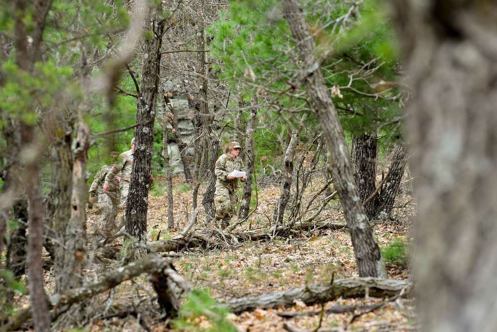 Two individuals in military camouflage uniforms are navigating through a wooded area with sparse undergrowth and fallen branches. One person appears to be holding a document or map, suggesting a land navigation or orienteering exercise. The image is taken from a distance through tree cover, partially obscuring the subjects.