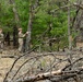 Two individuals in military camouflage uniforms are standing in a wooded area, appearing to examine or discuss something near a tree. The forest setting includes a mix of pine trees and deciduous trees, with fallen branches and dry leaves covering the ground in the foreground. The scene suggests a field training exercise or environmental assessment activity.