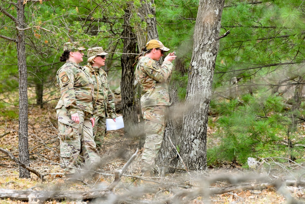 Three military personnel in OCP (Operational Camouflage Pattern) uniforms are conducting what appears to be a land navigation or orienteering exercise in a wooded area. One individual is using a compass or similar navigation instrument while the others observe and hold what appears to be a map or document. The setting is a pine forest with sparse undergrowth and leaf-covered ground.