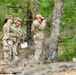 Three military personnel in OCP (Operational Camouflage Pattern) uniforms are conducting what appears to be a land navigation or orienteering exercise in a wooded area. One individual is using a compass or similar navigation instrument while the others observe and hold what appears to be a map or document. The setting is a pine forest with sparse undergrowth and leaf-covered ground.