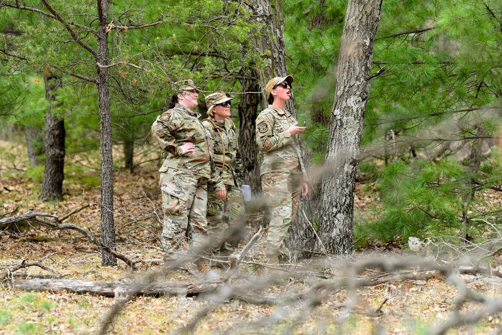 Three military personnel in OCP (Operational Camouflage Pattern) uniforms are standing in a wooded area, appearing to observe or assess something above them. One individual on the right appears to be speaking or gesturing while the others look upward. The setting suggests a field training exercise or outdoor assessment activity.