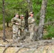 Three military personnel in OCP (Operational Camouflage Pattern) uniforms are standing in a wooded area, appearing to observe or assess something above them. One individual on the right appears to be speaking or gesturing while the others look upward. The setting suggests a field training exercise or outdoor assessment activity.