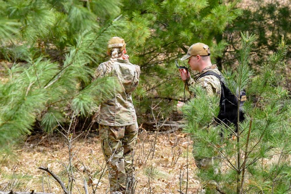 Two military personnel in OCP (Operational Camouflage Pattern) uniforms are operating in a wooded area with pine trees. The individual on the left, identified by a name tape reading 'HORNE,' appears to be observing or scanning the treeline, while the individual on the right is using a handheld communication device or navigation tool. The setting suggests a field training exercise or operational patrol activity.