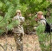 Two military personnel in OCP (Operational Camouflage Pattern) uniforms are operating in a wooded area with pine trees. The individual on the left, identified by a name tape reading 'HORNE,' appears to be observing or scanning the treeline, while the individual on the right is using a handheld communication device or navigation tool. The setting suggests a field training exercise or operational patrol activity.