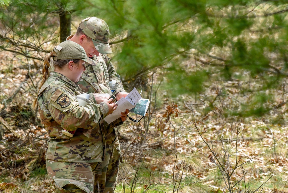 Two military personnel in MultiCam/OCP camouflage uniforms are standing in a wooded area reviewing a map or document together. Both wear matching patrol caps and unit patches on their left shoulders, suggesting they are engaged in a land navigation or field training exercise. The setting appears to be a temperate forest with pine trees and leaf-covered ground.