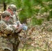 Two military personnel in MultiCam/OCP camouflage uniforms are standing in a wooded area reviewing a map or document together. Both wear matching patrol caps and unit patches on their left shoulders, suggesting they are engaged in a land navigation or field training exercise. The setting appears to be a temperate forest with pine trees and leaf-covered ground.
