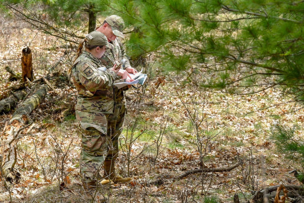Two military personnel in OCP (Operational Camouflage Pattern) uniforms are standing in a wooded area reviewing documents or a map together. They appear to be conducting land navigation or a field training exercise, consulting printed materials in an outdoor forest environment. The setting shows a sparse pine forest with leaf-covered ground during what appears to be early spring or late winter.