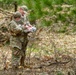 Two military personnel in OCP (Operational Camouflage Pattern) uniforms are standing in a wooded area reviewing documents or a map together. They appear to be conducting land navigation or a field training exercise, consulting printed materials in an outdoor forest environment. The setting shows a sparse pine forest with leaf-covered ground during what appears to be early spring or late winter.