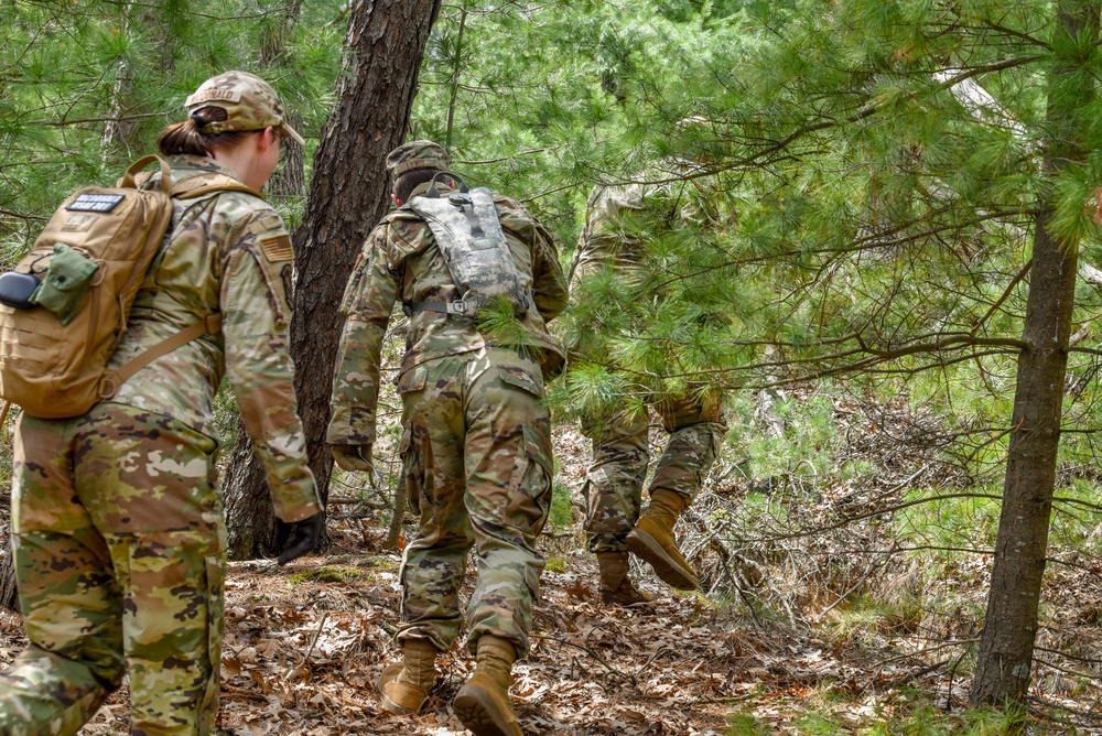 Three military personnel in camouflage uniforms are moving through a wooded forest terrain, navigating uphill through pine trees and leaf-covered ground. They are equipped with tactical gear, backpacks, and combat boots, suggesting a field training exercise or patrol movement. The lead soldier on the left appears to be female based on visible hair and body profile.