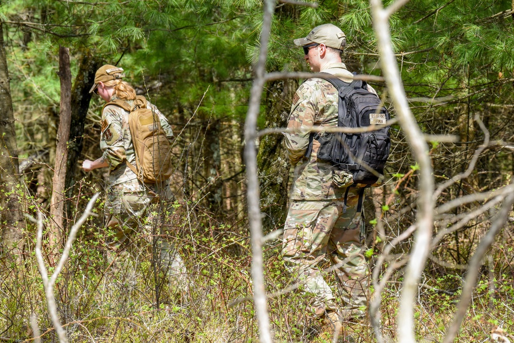 Two military personnel in OCP (Operational Camouflage Pattern) uniforms are moving through a wooded area carrying tactical backpacks. They appear to be conducting a land navigation or field training exercise in a forested environment with mixed pine and deciduous trees. The image is taken from a partially obscured angle through bare branches, suggesting early spring conditions.