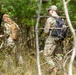 Two military personnel in OCP (Operational Camouflage Pattern) uniforms are moving through a wooded area carrying tactical backpacks. They appear to be conducting a land navigation or field training exercise in a forested environment with mixed pine and deciduous trees. The image is taken from a partially obscured angle through bare branches, suggesting early spring conditions.