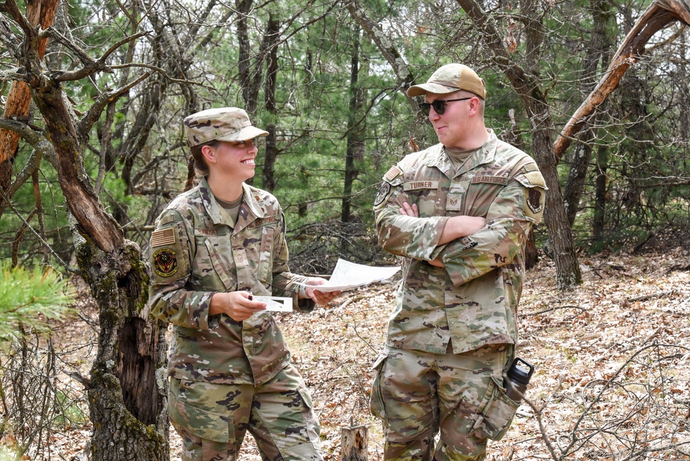 Two U.S. Air Force personnel in OCP (Operational Camouflage Pattern) uniforms are conversing in a wooded outdoor setting. The female airman on the left is holding documents and smiling, while the male airman on the right, identified by his name tape as 'Turner,' stands with arms crossed wearing sunglasses. They appear to be engaged in a field training exercise or outdoor operational activity.