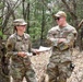 Two U.S. Air Force personnel in OCP (Operational Camouflage Pattern) uniforms are conversing in a wooded outdoor setting. The female airman on the left is holding documents and smiling, while the male airman on the right, identified by his name tape as 'Turner,' stands with arms crossed wearing sunglasses. They appear to be engaged in a field training exercise or outdoor operational activity.