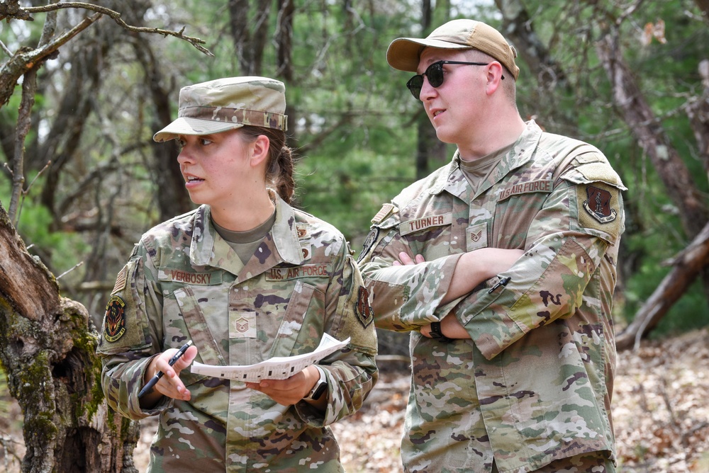 Two U.S. Air Force personnel in OCP (Operational Camouflage Pattern) uniforms are standing outdoors in a wooded area. The individual on the left, identified by name tape as 'VERBOSKY,' holds papers and a pen, while the individual on the right, identified as 'TURNER,' stands with arms crossed wearing sunglasses. Both appear to be engaged in a field training or assessment activity.