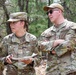 Two U.S. Air Force personnel in OCP (Operational Camouflage Pattern) uniforms are standing outdoors in a wooded area. The individual on the left, identified by name tape as 'VERBOSKY,' holds papers and a pen, while the individual on the right, identified as 'TURNER,' stands with arms crossed wearing sunglasses. Both appear to be engaged in a field training or assessment activity.