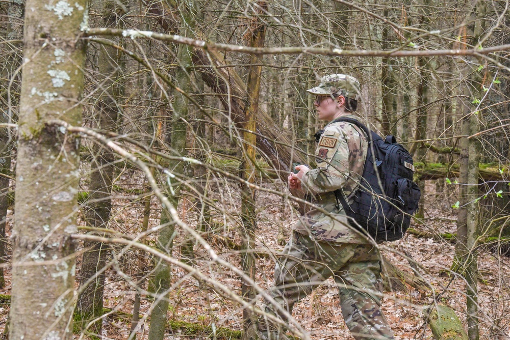A soldier in MultiCam/OCP camouflage uniform navigates through a deciduous forest with sparse undergrowth and leaf-covered ground. The individual is wearing a CBRN (Chemical, Biological, Radiological, and Nuclear) branch tab patch and carrying a black tactical backpack. The setting appears to be early spring based on the bare branches and minimal foliage.