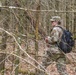 A soldier in MultiCam/OCP camouflage uniform navigates through a deciduous forest with sparse undergrowth and leaf-covered ground. The individual is wearing a CBRN (Chemical, Biological, Radiological, and Nuclear) branch tab patch and carrying a black tactical backpack. The setting appears to be early spring based on the bare branches and minimal foliage.