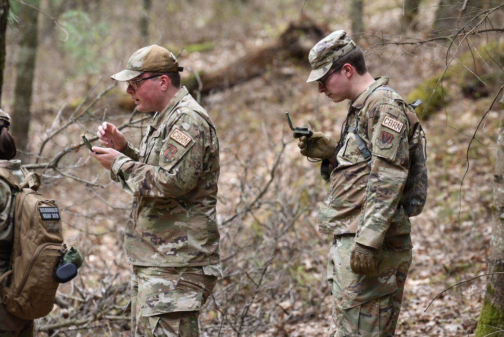 Two military personnel wearing OCP (Operational Camouflage Pattern) uniforms with 'CBRN' patches are standing in a wooded area, each examining handheld devices. A third individual is partially visible on the left side carrying a tactical backpack with a name tape reading 'McDONALD' and 'USAF 345'. The setting appears to be an outdoor field training exercise in a deciduous forest environment.