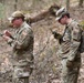 Two military personnel wearing OCP (Operational Camouflage Pattern) uniforms with 'CBRN' patches are standing in a wooded area, each examining handheld devices. A third individual is partially visible on the left side carrying a tactical backpack with a name tape reading 'McDONALD' and 'USAF 345'. The setting appears to be an outdoor field training exercise in a deciduous forest environment.