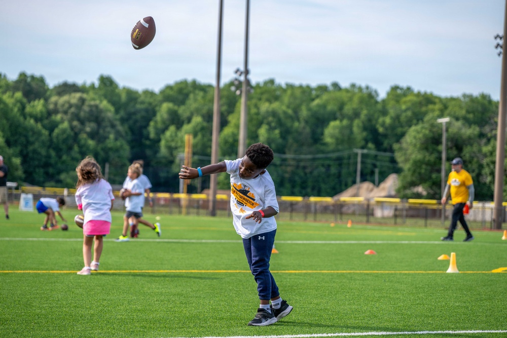 A youth football camp is taking place on an artificial turf field, with multiple children participating in drills. The focal point is a young boy in the foreground who has just thrown a football, captured mid-release with the ball visible in the air. Several other children and at least one adult coach are visible in the background engaged in various activities.