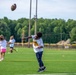 A youth football camp is taking place on an artificial turf field, with multiple children participating in drills. The focal point is a young boy in the foreground who has just thrown a football, captured mid-release with the ball visible in the air. Several other children and at least one adult coach are visible in the background engaged in various activities.