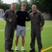 Three men are posing together on an athletic turf field. Two of the men are wearing olive/sage green military flight suits with patches, garrison caps, and name tags, consistent with U.S. Navy or Marine Corps aviator uniforms. The man in the center is wearing civilian athletic clothing — a black long-sleeve shirt and gray shorts.