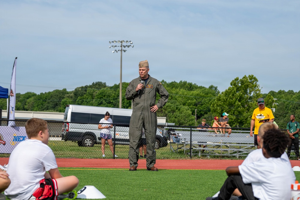 A military officer in an olive green flight suit and garrison cap is speaking into a microphone on a sports field, addressing a seated audience. The setting appears to be an outdoor athletic event or youth sports program, likely at a military installation based on the NEX (Navy Exchange) signage visible in the background. The speaker stands on artificial turf near a running track while various spectators and participants are visible in the background.