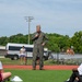 A military officer in an olive green flight suit and garrison cap is speaking into a microphone on a sports field, addressing a seated audience. The setting appears to be an outdoor athletic event or youth sports program, likely at a military installation based on the NEX (Navy Exchange) signage visible in the background. The speaker stands on artificial turf near a running track while various spectators and participants are visible in the background.