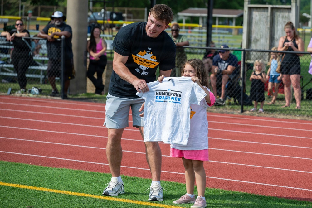 A tall young man wearing a black 'Eli Heidenreich' branded t-shirt is presenting a white 'Number One Draft Pick' t-shirt to a young smiling girl on a sports field with a red running track. The event appears to be a youth sports camp or community event, with spectators watching from behind a fence in the background. The girl is visibly excited and happy while holding up the shirt.