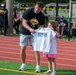 A tall young man wearing a black 'Eli Heidenreich' branded t-shirt is presenting a white 'Number One Draft Pick' t-shirt to a young smiling girl on a sports field with a red running track. The event appears to be a youth sports camp or community event, with spectators watching from behind a fence in the background. The girl is visibly excited and happy while holding up the shirt.