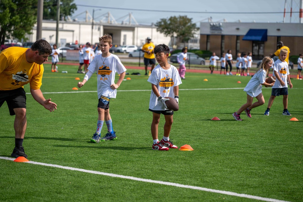 A youth football camp is taking place on an artificial turf field, with children in white t-shirts participating in drills supervised by adult coaches in yellow shirts. One child in the foreground is holding a football while standing near orange cone markers, appearing to receive instruction from a coach. Multiple groups of children are visible in the background engaged in similar football activities.
