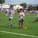 A youth football camp is taking place on an artificial turf field, with children in white t-shirts participating in drills supervised by adult coaches in yellow shirts. One child in the foreground is holding a football while standing near orange cone markers, appearing to receive instruction from a coach. Multiple groups of children are visible in the background engaged in similar football activities.