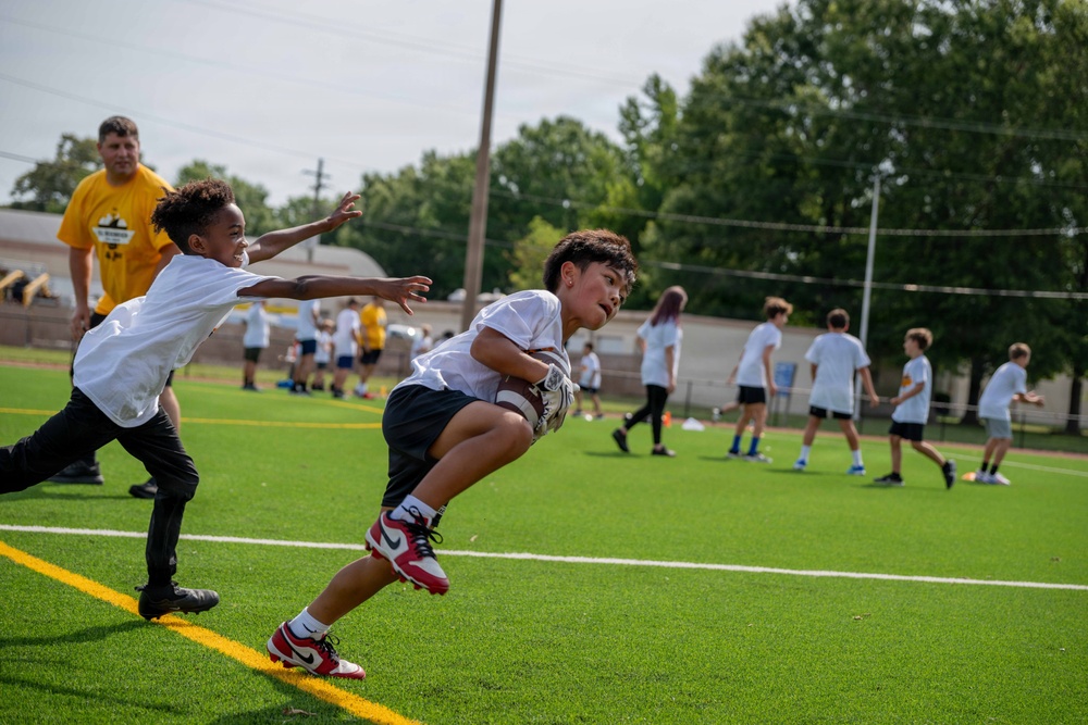 A youth football camp or practice session is taking place on an artificial turf field. In the foreground, a child carrying a football attempts to evade a defender who is reaching out to grab a flag or make a tag. Multiple other children and at least one adult coach in a yellow shirt are visible in the background engaged in various drills.