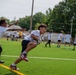 A youth football camp or practice session is taking place on an artificial turf field. In the foreground, a child carrying a football attempts to evade a defender who is reaching out to grab a flag or make a tag. Multiple other children and at least one adult coach in a yellow shirt are visible in the background engaged in various drills.