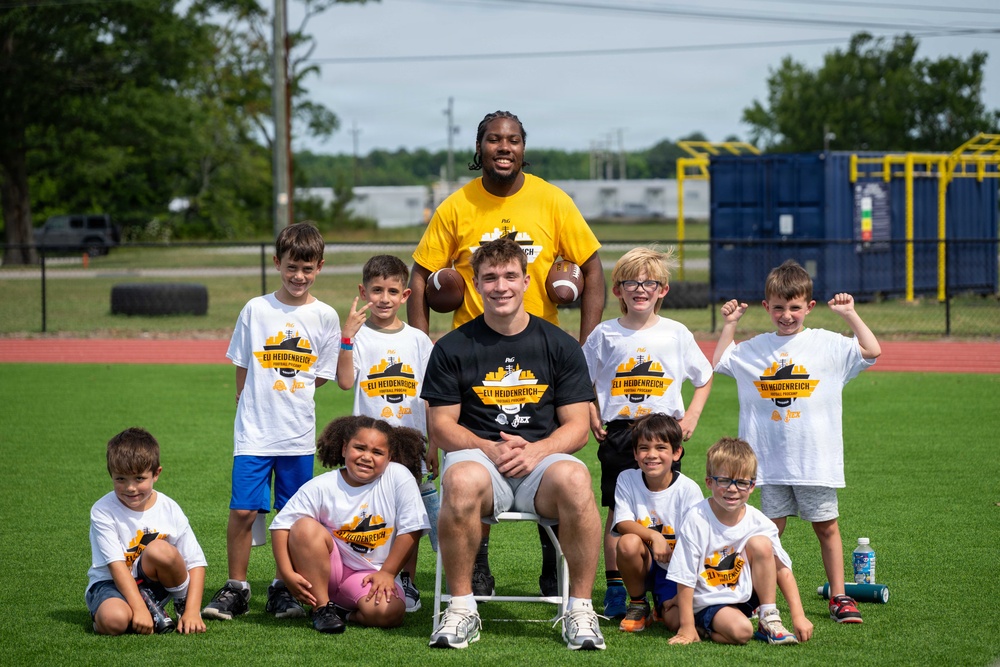 A group photo taken at the 'Eli Heidenreich Football Camp' on a green turf field. Two adult coaches — one wearing a black t-shirt with the camp logo and one in a yellow shirt — are posing with approximately nine young children, all wearing matching white camp t-shirts. The event appears to be a youth football camp with branding visible on the shirts.