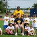 A group photo taken at the 'Eli Heidenreich Football Camp' on a green turf field. Two adult coaches — one wearing a black t-shirt with the camp logo and one in a yellow shirt — are posing with approximately nine young children, all wearing matching white camp t-shirts. The event appears to be a youth football camp with branding visible on the shirts.